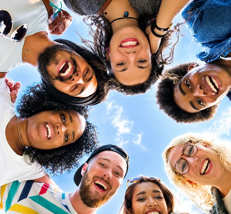 Grupo diverso de jóvenes sonriendo en círculo, vistos desde abajo contra el cielo